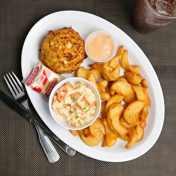 A crab cake platter with wedge fries, coleslaw, and dipping sauce on a white plate.