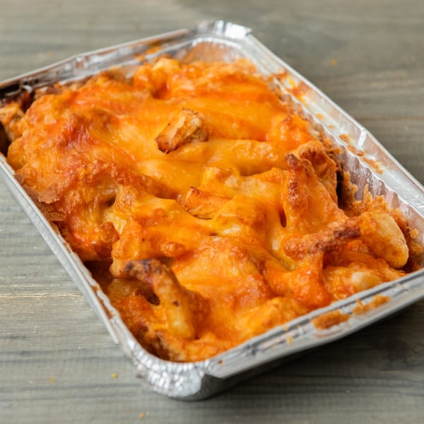 Cheesy baked pasta with chicken in a foil takeout container on a wooden table.