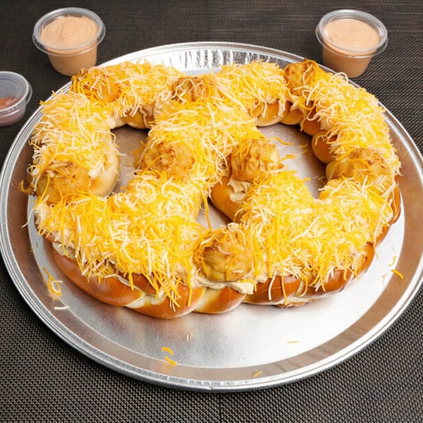 Large pretzel topped with shredded cheese and chicken, served with dipping sauces on a metal tray.