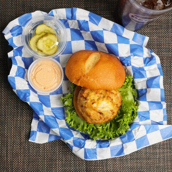 Crab cake sandwich on lettuce with pickles, sauce, and a drink on blue and white checkered paper.