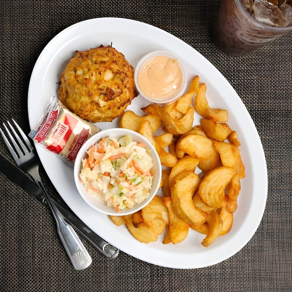A crab cake platter with coleslaw, potato wedges, and a side of sauce on a white plate.