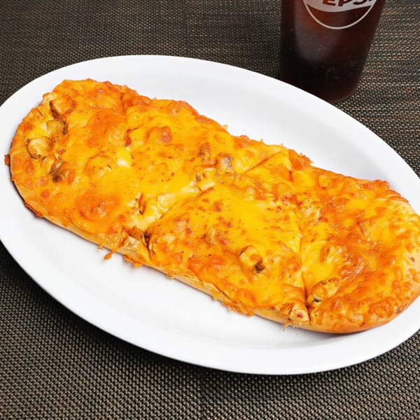 A long, oval flatbread pizza topped with melted orange cheese and chicken pieces, next to a Pepsi.
