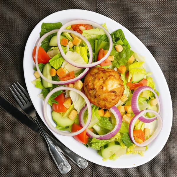 Overhead view of a fresh garden salad with a golden-brown crab cake, red onions, and croutons.